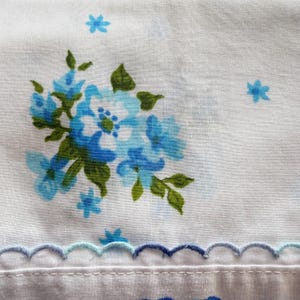 May include: White fabric with a floral pattern featuring blue flowers and green leaves. The design includes small blue flowers scattered across the fabric. A scalloped edge in shades of blue and aqua adds a decorative touch.