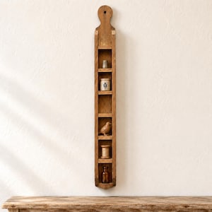 Op de afbeelding: Een hoge, smalle, verticale houten wandplank met zes open vakken. De plank heeft een afgeronde bovenkant met een ophanggat. In de vakken staan een metalen vingerhoed, een klein keramieken potje, een houten vogelbeeldje, een klosje en een glazen flesje.