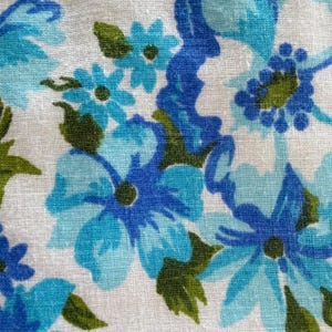 May include: Close-up of a fabric with a floral pattern. The design features blue and turquoise flowers with green leaves on a white background. The flowers vary in size and shape, creating a textured appearance.