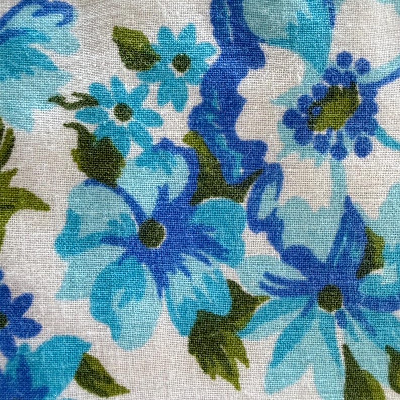 May include: Close-up of a fabric with a floral pattern. The design features blue and turquoise flowers with green leaves on a white background. The flowers vary in size and shape, creating a textured appearance.