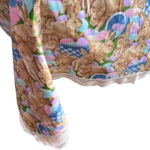 May include: A fabric with a repeating pattern of brown rabbits, Easter eggs, and baskets on a green, blue, and pink background. The fabric has a white lace trim along the edge. The design is likely for Easter or spring-themed crafts or home decor.