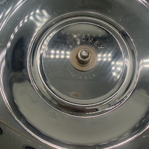 Op de afbeelding: Een cirkelvormig, gepolijst verchroomd metalen oppervlak met een centraal verhoogde naaf met een kleine metalen pin. De tekst "ROMEX" is bovenaan in het midden gegraveerd en de tekst "MADE IN U.S.A." is onderaan in de centrale naaf gegraveerd.