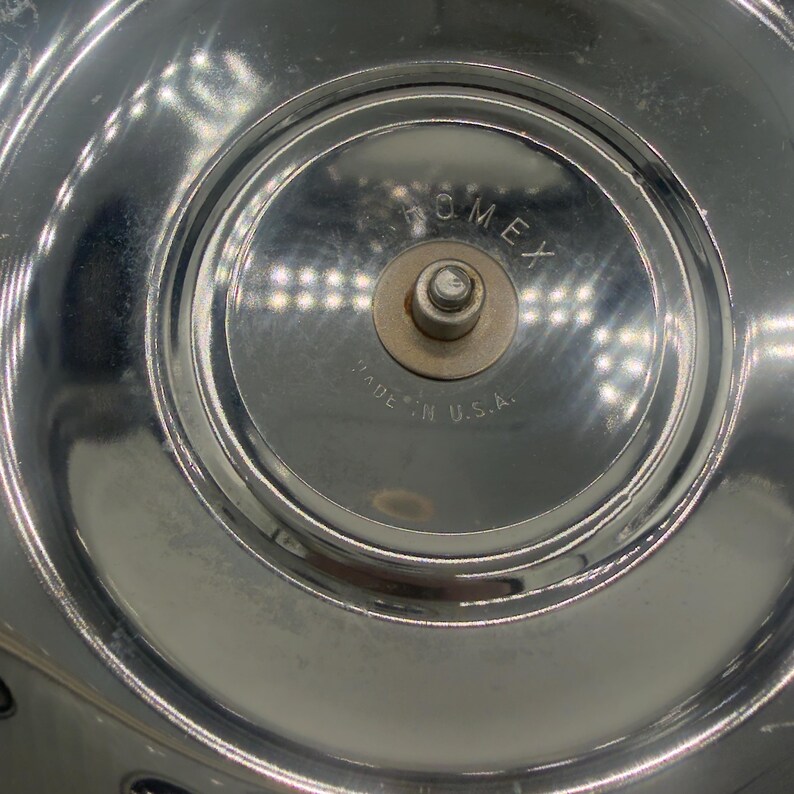 Op de afbeelding: Een cirkelvormig, gepolijst verchroomd metalen oppervlak met een centraal verhoogde naaf met een kleine metalen pin. De tekst "ROMEX" is bovenaan in het midden gegraveerd en de tekst "MADE IN U.S.A." is onderaan in de centrale naaf gegraveerd.