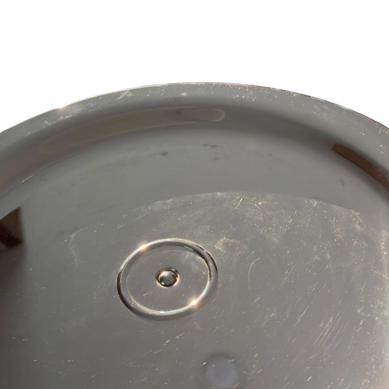 Op de afbeelding: Een cirkelvormig, reflecterend zilverkleurig metalen deksel met een glad, gepolijst oppervlak. Het midden van het deksel is gemarkeerd met een klein, verhoogd cirkelvormig detail met een minuscuul puntje in het midden voor een subtiel accent.