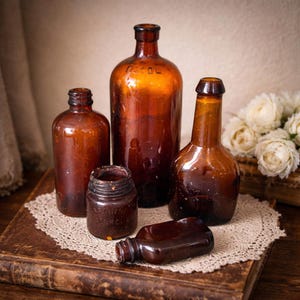 Könnte beinhalten: Eine Sammlung antiker brauner Glasflaschen in verschiedenen Größen. Die Flaschen sind auf einer Holzoberfläche mit einer Spitzendecke angeordnet. Die Flaschen haben eine Vintage-Ästhetik, einige weisen Gebrauchsspuren auf. Die Flaschen sind leer.