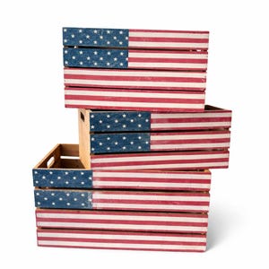 May include: A stack of wooden crates painted with the American flag design. The crates are painted with red and white stripes and a blue section with white stars. The crates are stacked on top of each other.