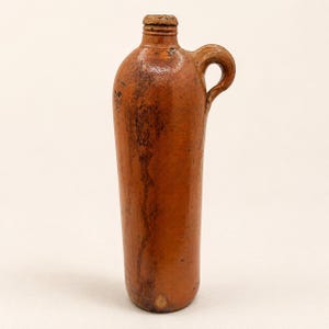 May include: A brown stoneware jug with a handle, featuring a narrow neck and a rounded body. The jug has a rustic appearance with visible imperfections and a glazed finish. The handle is arched, and the overall design suggests a vintage or antique style.