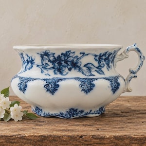 May include: A white ceramic chamber pot featuring a blue floral pattern around the upper rim and intricate blue scrollwork detailing along the base. The vessel includes a single ornate, curved handle on the right side and a wide, flared opening.