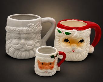 May include: Three ceramic mugs shaped like Santa Claus faces. One large white mug with a textured beard, one large mug with a red hat and painted facial features, and one small mug with a red handle and painted details on the face.