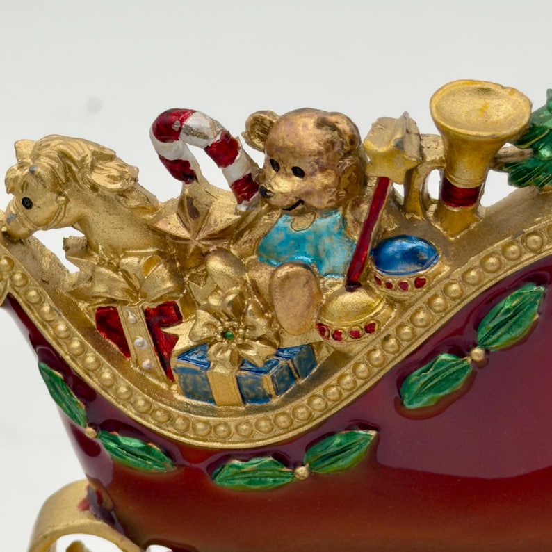May include: A decorative red sleigh figurine featuring a gold-toned rocking horse, a teddy bear in a blue shirt, a red and white candy cane, a gold trumpet, and wrapped gift boxes. The sleigh is accented with green holly leaves and gold trim.