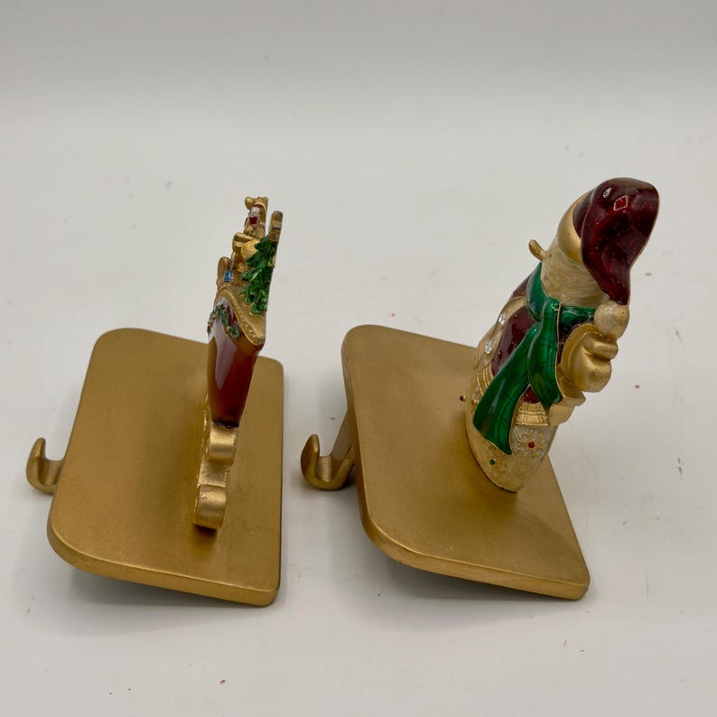 May include: Two gold-toned metal stocking holders with flat rectangular bases and curved hooks. One holder features a red stocking ornament with a green tree, while the other displays a snowman figure wearing a red hat and a green scarf.