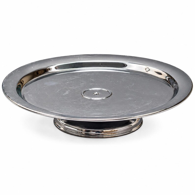 Op de afbeelding: Een ronde taartstandaard van zilverkleurig metaal met een gepolijst, reflecterend oppervlak. De standaard heeft een brede, platte schaal met een licht opstaande rand en een centrale cirkelvormige inkeping, rustend op een stevige voet.