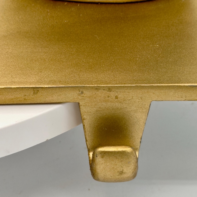 May include: A gold-colored metal stocking holder featuring a flat, rectangular base and a downward-facing hook. The surface has a smooth, matte finish with subtle variations in the metallic tone, designed to hang items from a flat edge or shelf.