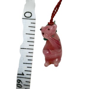 May include: A pink glass bear figurine with a green collar, suspended by a red cord. A white measuring tape is positioned vertically next to the bear, showing the figurine measures approximately 1.5 inches in total height from top to bottom.