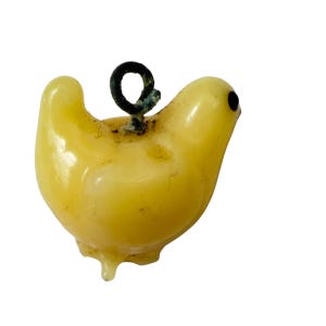 May include: A small, pale yellow bird-shaped charm made of a smooth, translucent material. The bird features a single dark, circular eye and a dark metal loop attached to the top of its back for hanging, showing signs of oxidation.