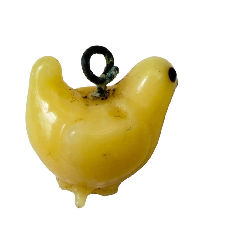 May include: A small, pale yellow bird-shaped charm made of a smooth, translucent material. The bird features a single dark, circular eye and a dark metal loop attached to the top of its back for hanging, showing signs of oxidation.