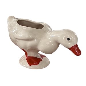 May include: A white ceramic duck-shaped planter with a red beak and red accents on the base. The planter has a hollowed-out area for holding plants or other items. The duck is in a downward-facing pose.