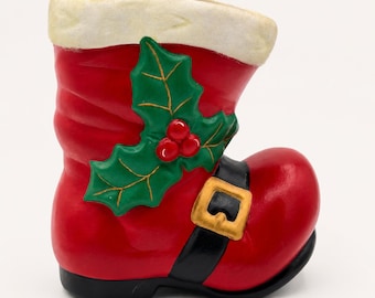 May include: A red ceramic boot-shaped container featuring a white textured cuff at the top, a black strap across the instep with a gold-colored buckle, and a green holly leaf decoration with three red berries attached to the side of the boot.