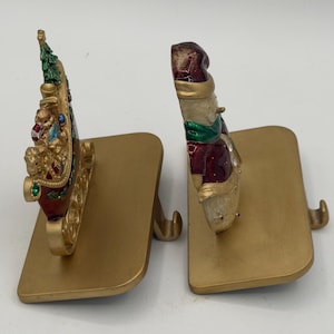 May include: Two gold-toned metal stocking holders. One features a red sleigh with a green Christmas tree and small figures, while the other depicts a snowman wearing a red hat and green scarf. Both have flat bases and a curved hook for hanging items.