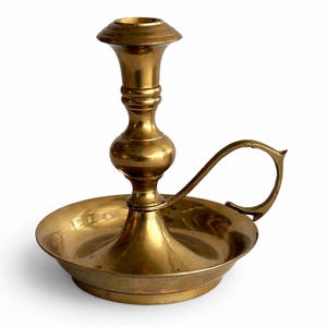 May include: A vintage brass candlestick holder. It features a round base, a central stem, and a handle. The candlestick holder has a classic design with a wide base and a cup-shaped top. The brass has a warm, golden colour. The height is approximately 15 cm.