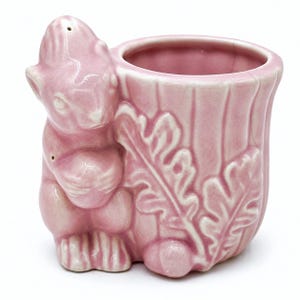 Puede incluir: Una maceta de cerámica rosa brillante con forma de tocón de árbol y una figura de ardilla adherida al lateral. La ardilla sostiene una bellota y el exterior de la maceta presenta detalles de hojas de roble en relieve y estrías verticales.