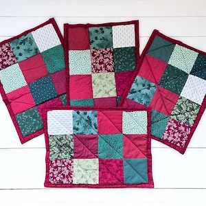 May include: Four handmade quilted placemats. Each placemat features a patchwork design with squares of red, green, and white fabric. The placemats are trimmed with a dark red border and are arranged on a white wooden surface.