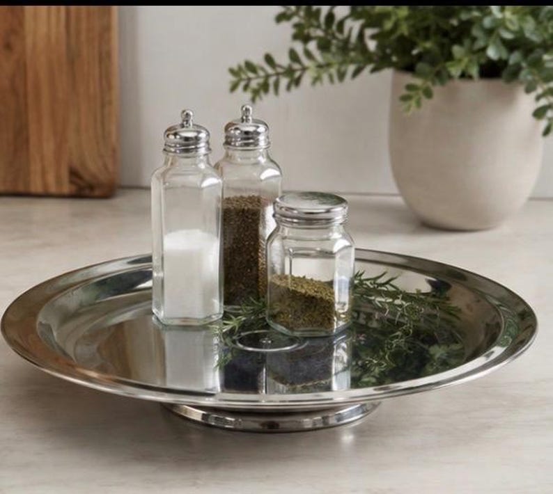 Op de afbeelding: Een ronde serveerschaal van gepolijst zilverkleurig metaal met drie doorzichtige glazen kruidenpotjes met zilverkleurige deksels. Twee hoge, zeshoekige potjes bevatten wit zout en zwarte peper, en een korter, rond potje bevat gedroogde groene kruiden.