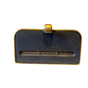May include: A rectangular desk accessory featuring a dark gray, textured cast iron base framed in a smooth gold-toned metal border. A horizontal slot runs across the center, revealing a gold-colored metal strip with two small, clear circular accents.