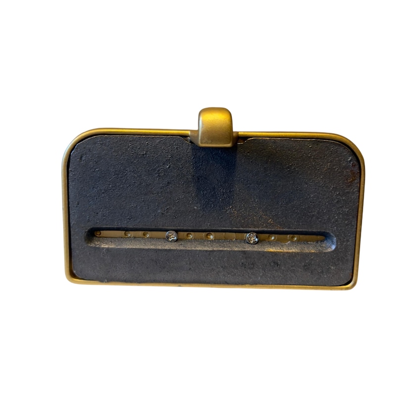 May include: A rectangular desk accessory featuring a dark gray, textured cast iron base framed in a smooth gold-toned metal border. A horizontal slot runs across the center, revealing a gold-colored metal strip with two small, clear circular accents.