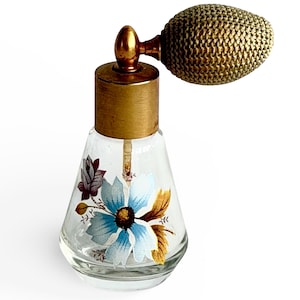 Puede incluir: Un frasco de perfume vintage con un cuerpo de vidrio transparente y un atomizador de latón. La botella presenta un diseño floral pintado a mano con detalles en azul y dorado. El atomizador tiene un diseño bulboso tejido.