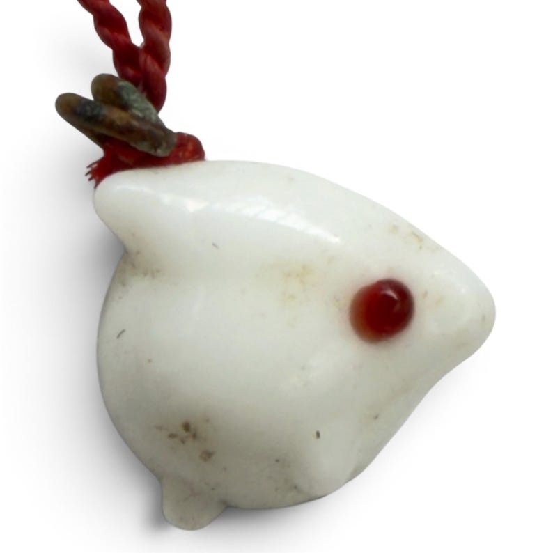 May include: A white ceramic pendant shaped like a small animal head with a single dark red eye, suspended from a twisted red cord. The pendant features a smooth, rounded surface with a small protrusion at the top and a metallic loop attachment.