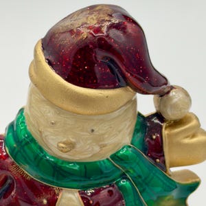 May include: A decorative figurine of a snowman wearing a deep red glittery hat with a gold band and a green scarf. The snowman has a cream-colored swirled finish, a small gold nose, and gold-toned accents on the hat and clothing details.