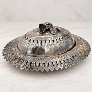 Könnte beinhalten: Eine ovale Servierschale aus silberfarbenem Metall mit abnehmbarem Deckel. Der Deckel hat einen blumenförmigen Griff und dekorative Ausschnitte. Der breite, flache Rand ist gewellt und mit detaillierten Blattgravuren verziert.