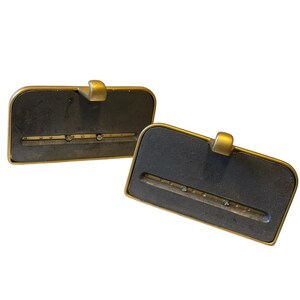 May include: Two rectangular bookends featuring a dark gray, textured central panel framed by a smooth, matte gold-toned metal border. Each piece includes a small, centered gold-toned hook at the top and a horizontal slot containing two small, clear crystals.