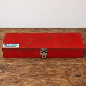 May include: A rectangular red metal tool box with a weathered finish and a single silver-colored metal latch on the front. A white rectangular label on the left side features the blue and white NAPA New Britain Power Tools logo and text.