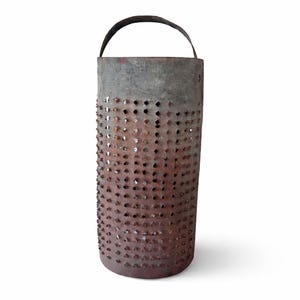May include: A cylindrical, rusty metal lantern with a handle. The lantern is perforated with numerous small holes, creating a textured surface. The metal has a weathered appearance, with shades of grey and brown.