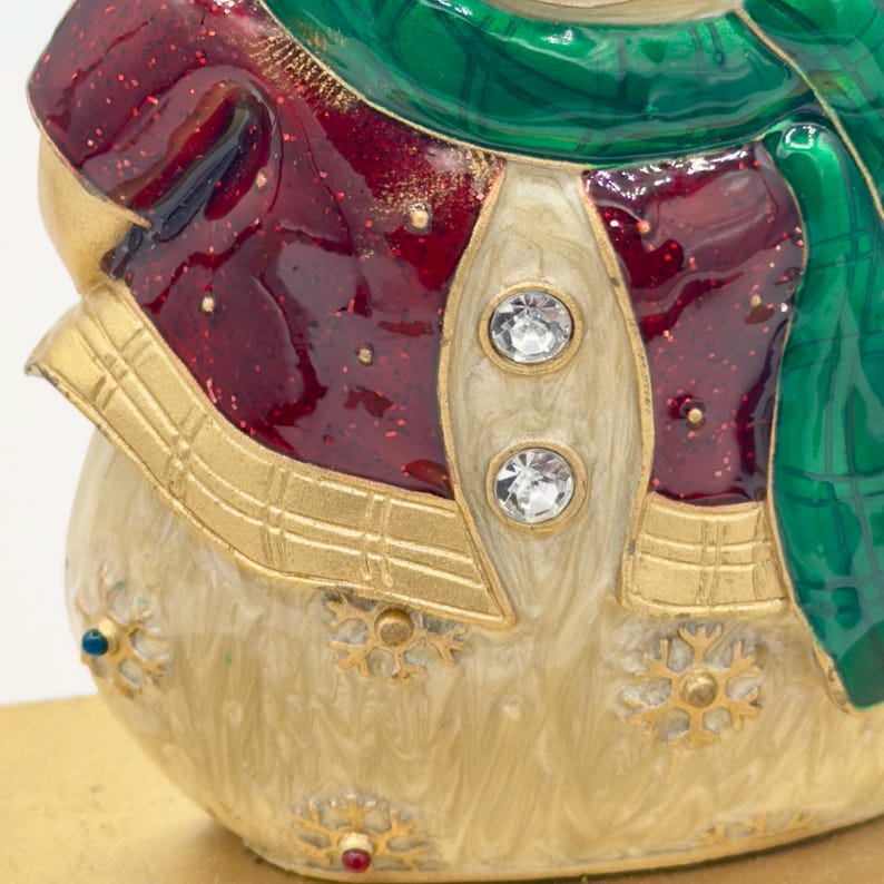 May include: A decorative snowman figurine featuring a cream-colored body with gold snowflake accents, a deep red jacket with glittery details, and a green scarf. The jacket is adorned with two clear round crystals as buttons.