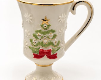 May include: A cream-colored ceramic mug with a pedestal base, featuring a raised, painted Christmas tree design with a gold star topper and a red bow at the base. The mug is decorated with embossed snowflake patterns and gold trim on the rim, handle, and base.