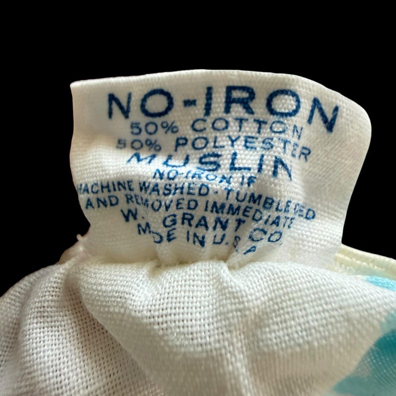 May include: Close-up of a white fabric tag with blue text. The tag reads "NO-IRON", "50% COTTON 50% POLYESTER MUSLIN", and other care instructions. The tag is from W. Grant Co. Made in U.S.A.