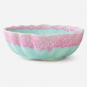 May include: A ceramic bowl with a scalloped rim. The bowl is a light turquoise color with a pink, dripped glaze around the rim. The bowl has a wavy, textured design on the exterior.