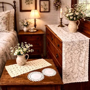May include: A bedroom scene featuring a wooden nightstand with a lamp, a bed with floral bedding, and a wooden dresser. A cream-colored lace table runner and matching placemats and coasters are on display, along with vases of white flowers.