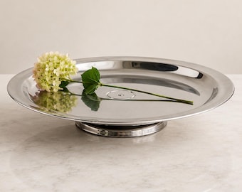 Può includere: Un vassoio a piedistallo in metallo color argento lucido con un bordo largo e piatto e una base circolare. Un singolo fiore di ortensia verde chiaro con un sottile stelo verde è posizionato orizzontalmente al centro della superficie liscia e riflettente.