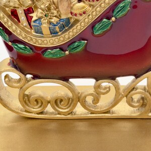 May include: A decorative sleigh figurine featuring a deep red body with gold scrollwork runners. The sleigh is filled with sculpted gift boxes, a gold bow, and green holly leaf accents. The finish is glossy with metallic gold detailing.