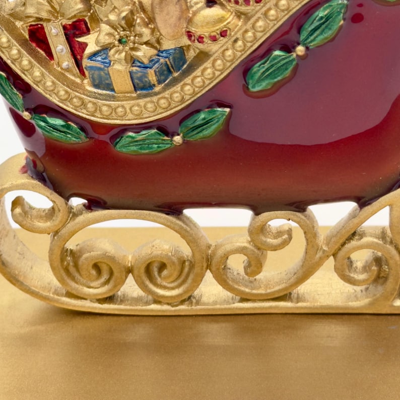 May include: A decorative sleigh figurine featuring a deep red body with gold scrollwork runners. The sleigh is filled with sculpted gift boxes, a gold bow, and green holly leaf accents. The finish is glossy with metallic gold detailing.