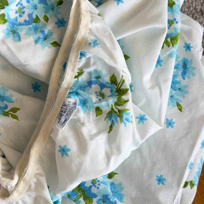 May include: White fitted sheet with a blue floral pattern and green leaves. The sheet has an elastic band and a tag that reads "Hurley Mattress Grand Tone". The floral design is repeated across the fabric.