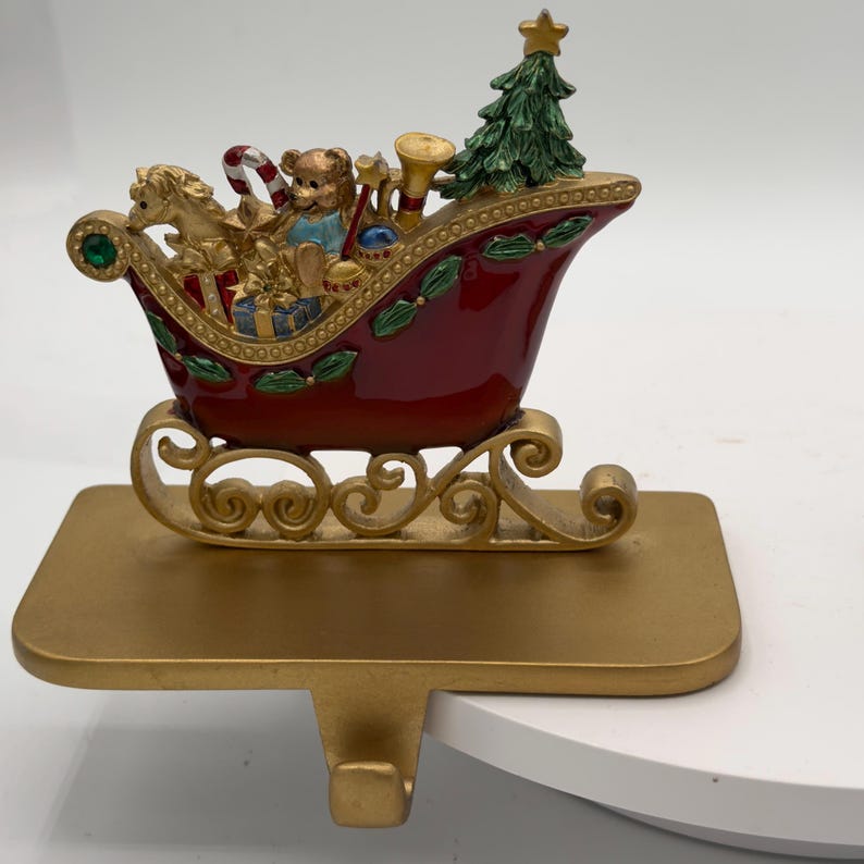May include: A decorative Christmas stocking holder featuring a deep red sleigh with gold scrollwork runners. The sleigh is filled with a miniature green Christmas tree, a teddy bear, a candy cane, a trumpet, and wrapped gift boxes.
