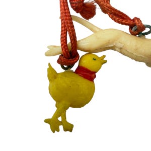 May include: A yellow chick charm with a red neck band hangs from an orange braided cord. The cord is looped around a pale, off-white, branch-like structure. The chick has a small beak, two legs, and is suspended by a metal jump ring.