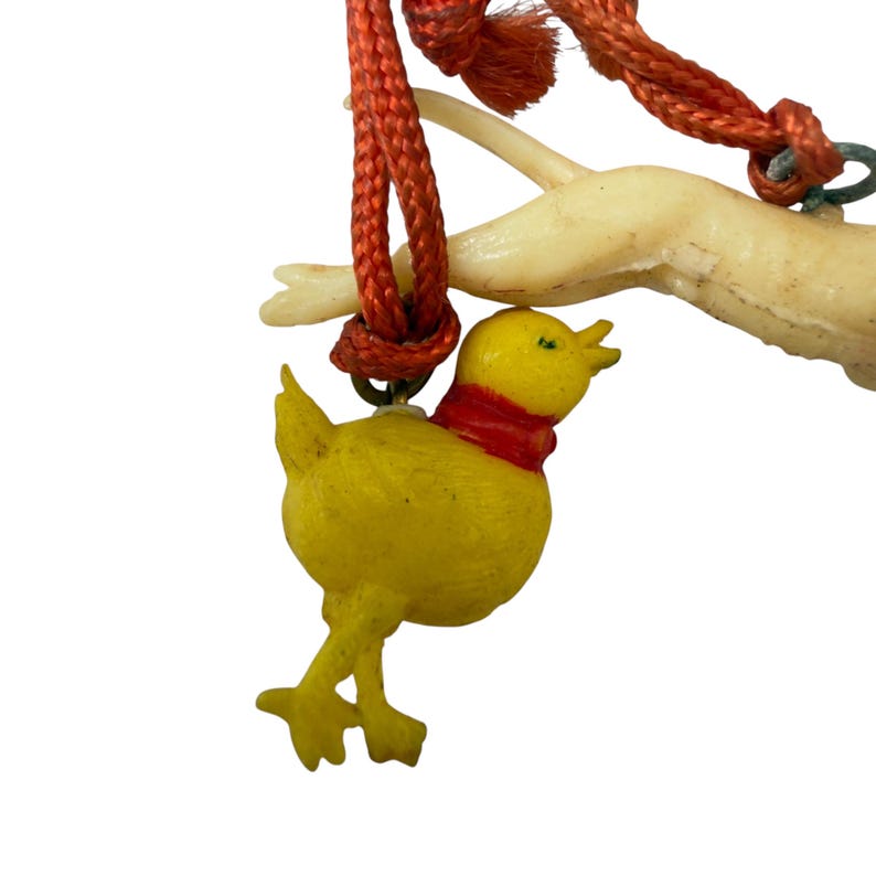 May include: A yellow chick charm with a red neck band hangs from an orange braided cord. The cord is looped around a pale, off-white, branch-like structure. The chick has a small beak, two legs, and is suspended by a metal jump ring.