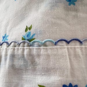 May include: Close-up of white fabric with blue floral patterns and a scalloped edge. The flowers are in shades of blue and green. The scalloped edge is a combination of light and dark blue thread, adding a decorative touch to the textile.