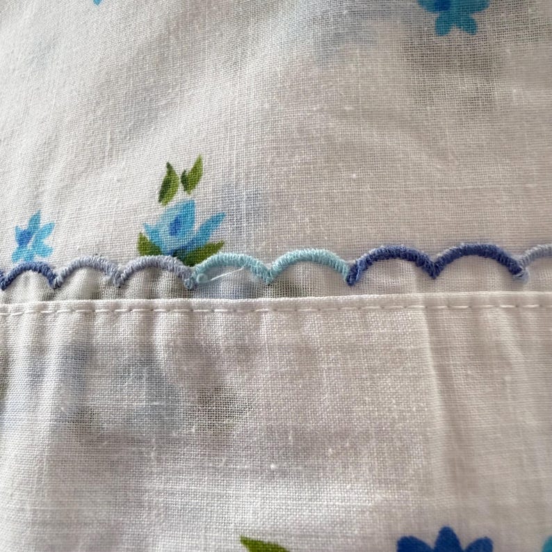 May include: Close-up of white fabric with blue floral patterns and a scalloped edge. The flowers are in shades of blue and green. The scalloped edge is a combination of light and dark blue thread, adding a decorative touch to the textile.